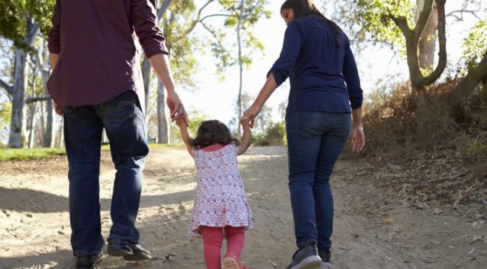 Adultos caminham ao ar livre de mãos dadas com uma criança, simbolizando acolhimento familiar, cuidado, proteção e convivência em ambiente seguro.