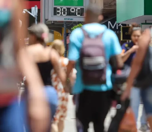 Semana terá calor intenso e chance de temporais em SC Pessoas caminham pela cidade em dia de calor em Santa Catarina, com termômetro de rua marcando 30°C ao fundo.
