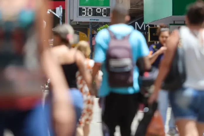 Pessoas caminham pela cidade em dia de calor em Santa Catarina, com termômetro de rua marcando 30°C ao fundo.