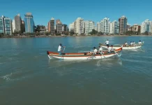 Canoas a remo durante a corrida de canoa a remo em Florianópolis, com equipes competindo na Beira-Mar Norte e prédios da cidade ao fundo.