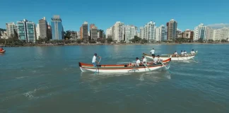 Canoas a remo durante a corrida de canoa a remo em Florianópolis, com equipes competindo na Beira-Mar Norte e prédios da cidade ao fundo.