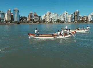 Canoas a remo durante a corrida de canoa a remo em Florianópolis, com equipes competindo na Beira-Mar Norte e prédios da cidade ao fundo.