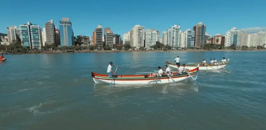Canoas a remo durante a corrida de canoa a remo em Florianópolis, com equipes competindo na Beira-Mar Norte e prédios da cidade ao fundo.