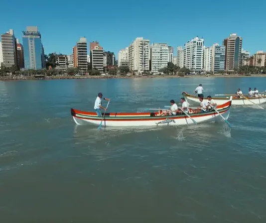 Canoas a remo durante a corrida de canoa a remo em Florianópolis, com equipes competindo na Beira-Mar Norte e prédios da cidade ao fundo.