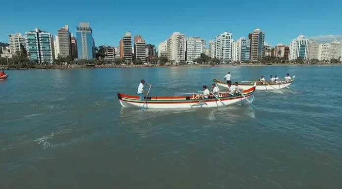 Canoas a remo durante a corrida de canoa a remo em Florianópolis, com equipes competindo na Beira-Mar Norte e prédios da cidade ao fundo.