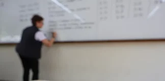 Professor escreve em quadro em sala de aula enquanto, em primeiro plano, aparece material da Secretaria de Educação de Santa Catarina, ilustrando o contexto do concurso público da rede estadual de ensino.