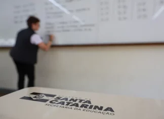 SC lança concurso público da Educação com até 10 mil vagas para quadro efetivo Professor escreve em quadro em sala de aula enquanto, em primeiro plano, aparece material da Secretaria de Educação de Santa Catarina, ilustrando o contexto do concurso público da rede estadual de ensino.