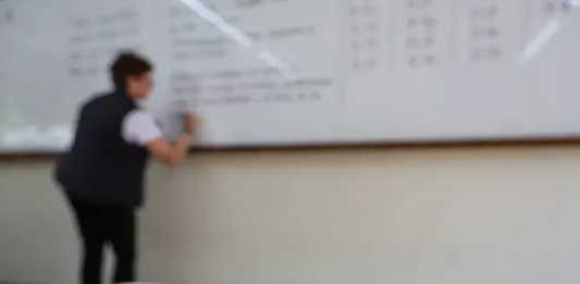 Professor escreve em quadro em sala de aula enquanto, em primeiro plano, aparece material da Secretaria de Educação de Santa Catarina, ilustrando o contexto do concurso público da rede estadual de ensino.