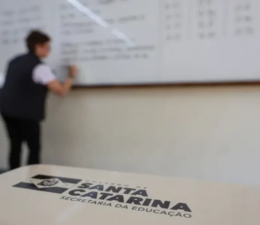 SC lança concurso público da Educação com até 10 mil vagas para quadro efetivo Professor escreve em quadro em sala de aula enquanto, em primeiro plano, aparece material da Secretaria de Educação de Santa Catarina, ilustrando o contexto do concurso público da rede estadual de ensino.