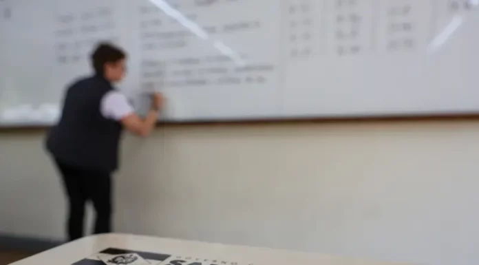 Professor escreve em quadro em sala de aula enquanto, em primeiro plano, aparece material da Secretaria de Educação de Santa Catarina, ilustrando o contexto do concurso público da rede estadual de ensino.