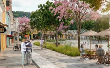 Imagem ilustrativa apresenta proposta de requalificação do Centro de Florianópolis, com calçadas amplas, ciclovia, árvores floridas e praça com fonte e mesas ao ar livre. A cena mostra pessoas caminhando, pedalando e convivendo no espaço público, ilustrando a proposta de planejamento urbano que prioriza pedestres, áreas verdes e convivência no projeto Floripa Centro.