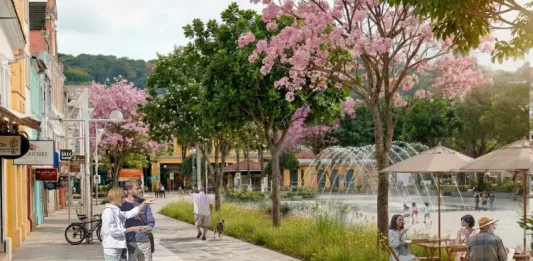 Imagem ilustrativa apresenta proposta de requalificação do Centro de Florianópolis, com calçadas amplas, ciclovia, árvores floridas e praça com fonte e mesas ao ar livre. A cena mostra pessoas caminhando, pedalando e convivendo no espaço público, ilustrando a proposta de planejamento urbano que prioriza pedestres, áreas verdes e convivência no projeto Floripa Centro.
