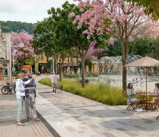 Imagem ilustrativa apresenta proposta de requalificação do Centro de Florianópolis, com calçadas amplas, ciclovia, árvores floridas e praça com fonte e mesas ao ar livre. A cena mostra pessoas caminhando, pedalando e convivendo no espaço público, ilustrando a proposta de planejamento urbano que prioriza pedestres, áreas verdes e convivência no projeto Floripa Centro.