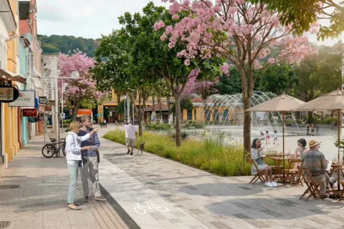 Imagem ilustrativa apresenta proposta de requalificação do Centro de Florianópolis, com calçadas amplas, ciclovia, árvores floridas e praça com fonte e mesas ao ar livre. A cena mostra pessoas caminhando, pedalando e convivendo no espaço público, ilustrando a proposta de planejamento urbano que prioriza pedestres, áreas verdes e convivência no projeto Floripa Centro.
