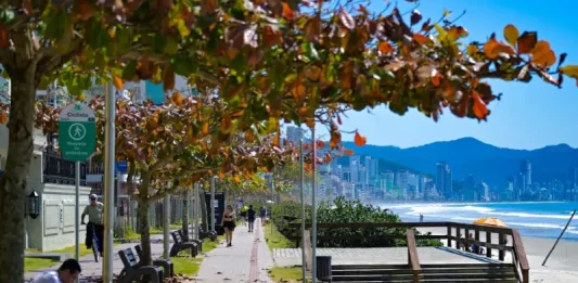 Outono em Santa Catarina terá calor acima da média e variação térmica Imagem de calçadão à beira-mar em Santa Catarina, com pessoas caminhando e céu ensolarado, utilizada para ilustrar a reportagem sobre o início do outono com temperaturas acima da média, dias ainda quentes, variações térmicas ao longo do dia e a presença de instabilidades típicas da estação.