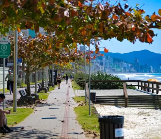 Imagem de calçadão à beira-mar em Santa Catarina, com pessoas caminhando e céu ensolarado, utilizada para ilustrar a reportagem sobre o início do outono com temperaturas acima da média, dias ainda quentes, variações térmicas ao longo do dia e a presença de instabilidades típicas da estação.