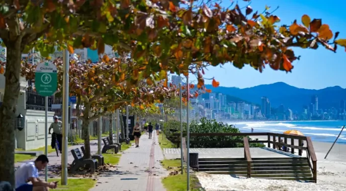 Outono em Santa Catarina terá calor acima da média e variação térmica Imagem de calçadão à beira-mar em Santa Catarina, com pessoas caminhando e céu ensolarado, utilizada para ilustrar a reportagem sobre o início do outono com temperaturas acima da média, dias ainda quentes, variações térmicas ao longo do dia e a presença de instabilidades típicas da estação.