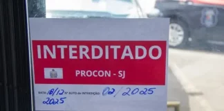 Placa de ‘interditado pelo Procon de São José’ colada na porta de vidro de um comércio, representando suspensão de alvará em São José.