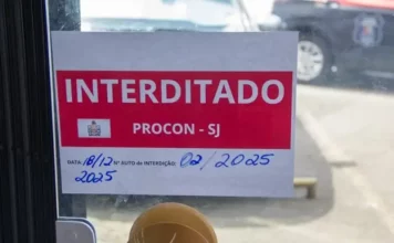 Placa de ‘interditado pelo Procon de São José’ colada na porta de vidro de um comércio, representando suspensão de alvará em São José.