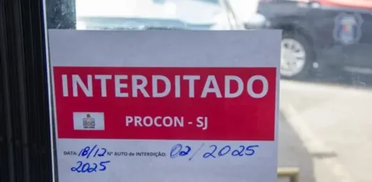 Placa de ‘interditado pelo Procon de São José’ colada na porta de vidro de um comércio, representando suspensão de alvará em São José.