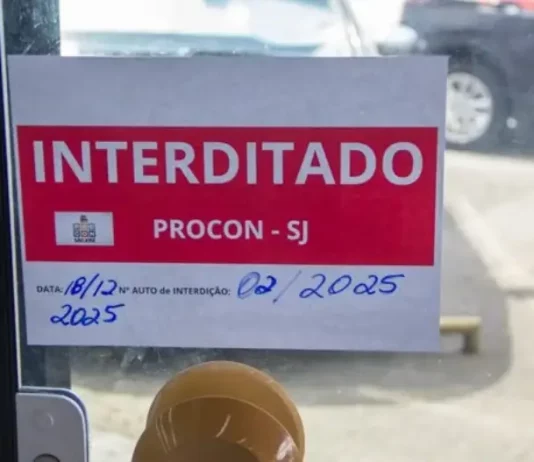 Placa de ‘interditado pelo Procon de São José’ colada na porta de vidro de um comércio, representando suspensão de alvará em São José.