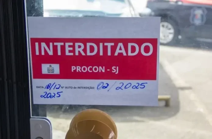 Placa de ‘interditado pelo Procon de São José’ colada na porta de vidro de um comércio, representando suspensão de alvará em São José.