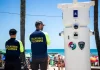 Voluntários de segurança em Florianópolis observam movimento na praia ao lado de torre de monitoramento com símbolos da Guarda Municipal e da Polícia Militar.