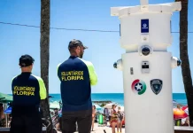 Voluntários de segurança em Florianópolis observam movimento na praia ao lado de torre de monitoramento com símbolos da Guarda Municipal e da Polícia Militar.