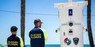 Voluntários de segurança em Florianópolis observam movimento na praia ao lado de torre de monitoramento com símbolos da Guarda Municipal e da Polícia Militar.