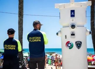 Voluntários de segurança em Florianópolis observam movimento na praia ao lado de torre de monitoramento com símbolos da Guarda Municipal e da Polícia Militar.
