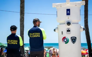 Voluntários de segurança em Florianópolis observam movimento na praia ao lado de torre de monitoramento com símbolos da Guarda Municipal e da Polícia Militar.