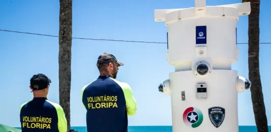 Voluntários de segurança em Florianópolis observam movimento na praia ao lado de torre de monitoramento com símbolos da Guarda Municipal e da Polícia Militar.