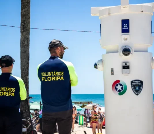 Voluntários de segurança em Florianópolis observam movimento na praia ao lado de torre de monitoramento com símbolos da Guarda Municipal e da Polícia Militar.