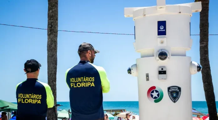 Voluntários de segurança em Florianópolis observam movimento na praia ao lado de torre de monitoramento com símbolos da Guarda Municipal e da Polícia Militar.
