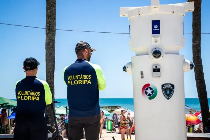 Voluntários de segurança em Florianópolis observam movimento na praia ao lado de torre de monitoramento com símbolos da Guarda Municipal e da Polícia Militar.