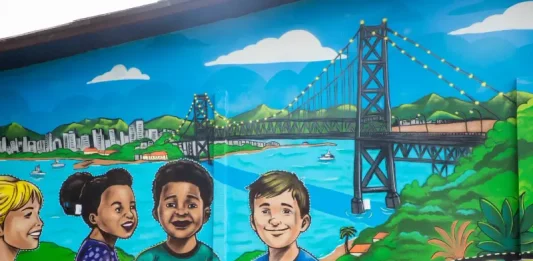 Crianças participam de criação de mural no Jardim Botânico em homenagem à Ponte Hercílio Luz Mural de grafite no Jardim Botânico de Florianópolis retrata crianças e a Ponte Hercílio Luz, simbolizando a participação infantil em projeto de arte e cidadania.