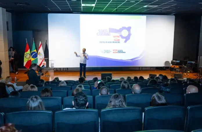 Governador Jorginho Mello durante apresentação no lançamento do Santa Catarina Canta 2026 em auditório, com público presente, destacando a abertura das inscrições e a divulgação do festival de música no estado.