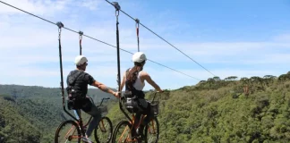 Duas pessoas praticam cicloturismo em cabo suspenso (skybike) sobre área de mata, ilustrando atividade de turismo de aventura com vista panorâmica e contato com a natureza.