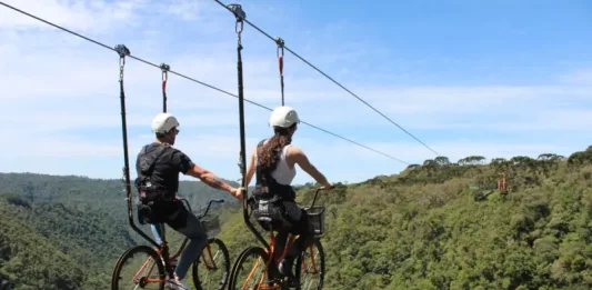 Duas pessoas praticam cicloturismo em cabo suspenso (skybike) sobre área de mata, ilustrando atividade de turismo de aventura com vista panorâmica e contato com a natureza.