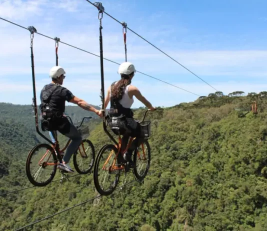 Duas pessoas praticam cicloturismo em cabo suspenso (skybike) sobre área de mata, ilustrando atividade de turismo de aventura com vista panorâmica e contato com a natureza.