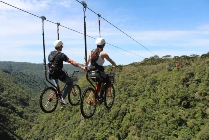 Duas pessoas praticam cicloturismo em cabo suspenso (skybike) sobre área de mata, ilustrando atividade de turismo de aventura com vista panorâmica e contato com a natureza.