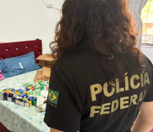 Policial federal durante ação contra comércio ilegal de medicamentos, com remédios apreendidos sobre cama em residência.