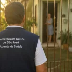 Golpistas se passam por agentes de saúde para aplicar fraudes em São José Imagem gerada por IA mostra um homem com colete de agente de saúde da Secretaria de São José em frente a uma residência, ilustrando a ação de golpistas que se passam por profissionais para aplicar fraudes.