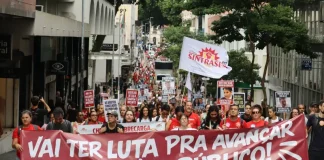 Servidores públicos participam de manifestação durante a greve servidores Florianópolis, com faixa do Sintrasem e cartazes com reivindicações da categoria em rua central.