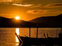 Imagem de um pôr do sol em Santa Catarina, com o sol refletindo sobre a água e silhuetas de barcos em destaque. A cena transmite céu limpo e clima quente, ilustrando o tempo firme e ensolarado previsto para o feriado de Tiradentes.