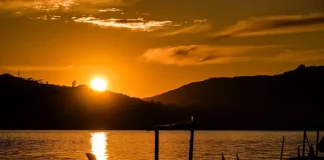 Imagem de um pôr do sol em Santa Catarina, com o sol refletindo sobre a água e silhuetas de barcos em destaque. A cena transmite céu limpo e clima quente, ilustrando o tempo firme e ensolarado previsto para o feriado de Tiradentes.