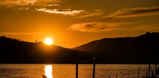 Imagem de um pôr do sol em Santa Catarina, com o sol refletindo sobre a água e silhuetas de barcos em destaque. A cena transmite céu limpo e clima quente, ilustrando o tempo firme e ensolarado previsto para o feriado de Tiradentes.