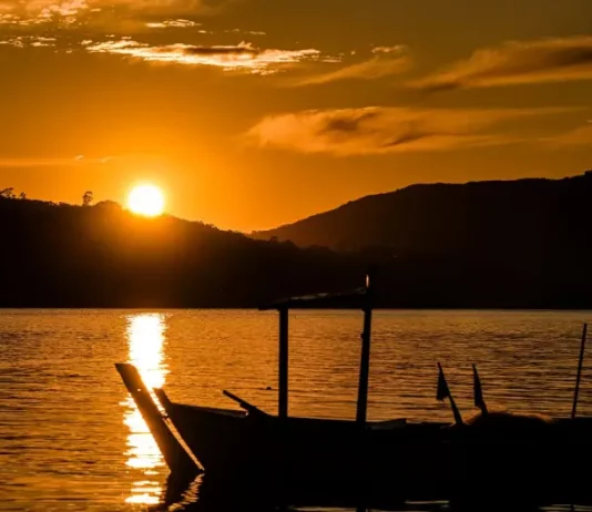 Imagem de um pôr do sol em Santa Catarina, com o sol refletindo sobre a água e silhuetas de barcos em destaque. A cena transmite céu limpo e clima quente, ilustrando o tempo firme e ensolarado previsto para o feriado de Tiradentes.