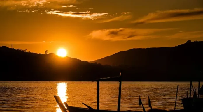 Imagem de um pôr do sol em Santa Catarina, com o sol refletindo sobre a água e silhuetas de barcos em destaque. A cena transmite céu limpo e clima quente, ilustrando o tempo firme e ensolarado previsto para o feriado de Tiradentes.