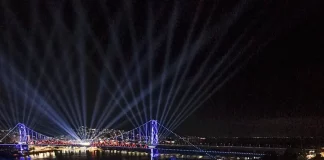 Vista aérea mostra o público reunido na Beira-Mar Continental durante as comemorações do centenário da Ponte Hercílio Luz, em Florianópolis, com palco de shows, iluminação especial e feixes de luz sobre a ponte durante evento noturno.